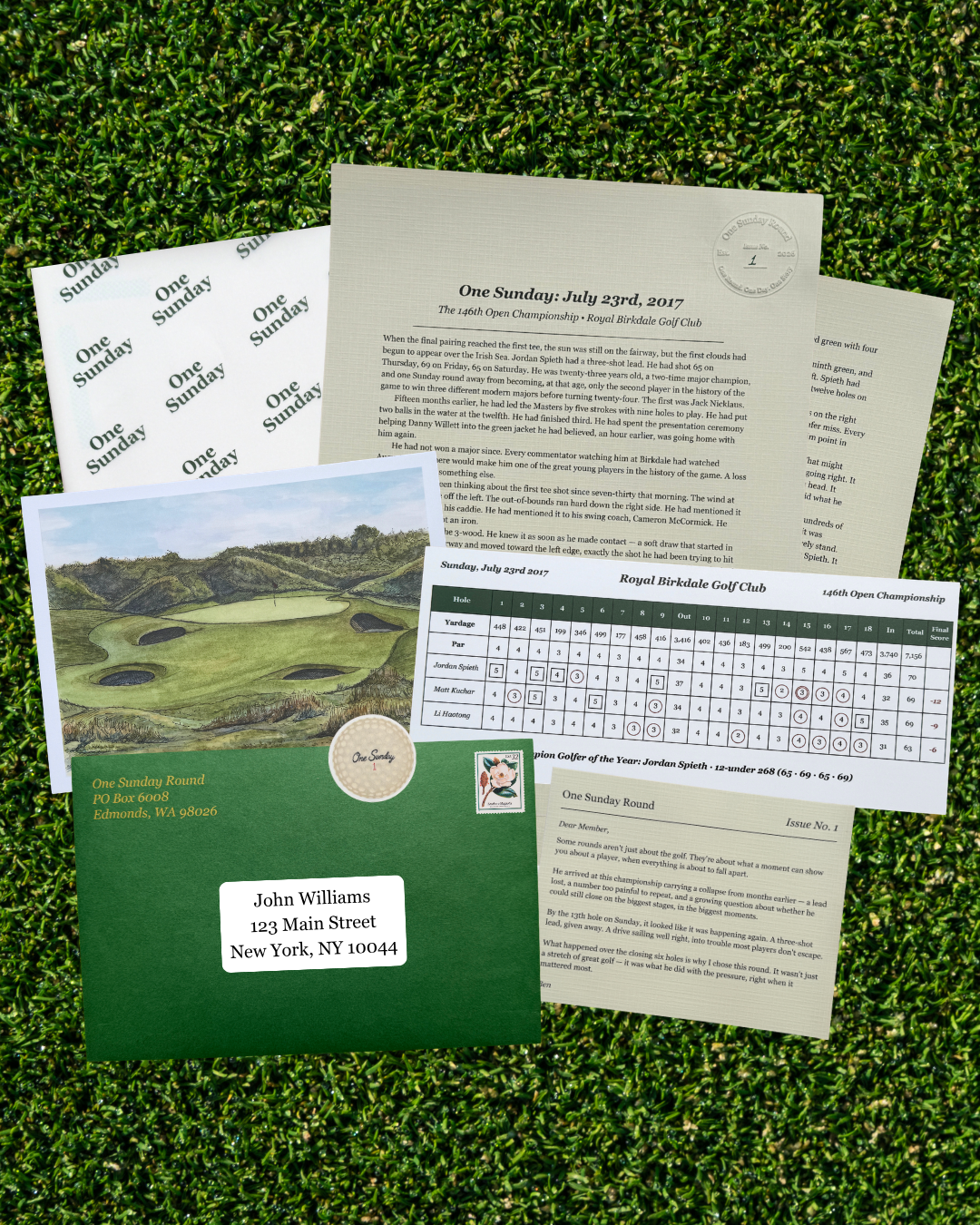 A golf-themed stationery set arranged on artificial grass, including a green envelope with a printed golf course photo, cream letterhead papers, a round-robin sheet, and a name label sticker.